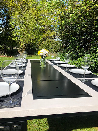 Outdoor dining setup with a black cooktop on a table, surrounded by greenery.