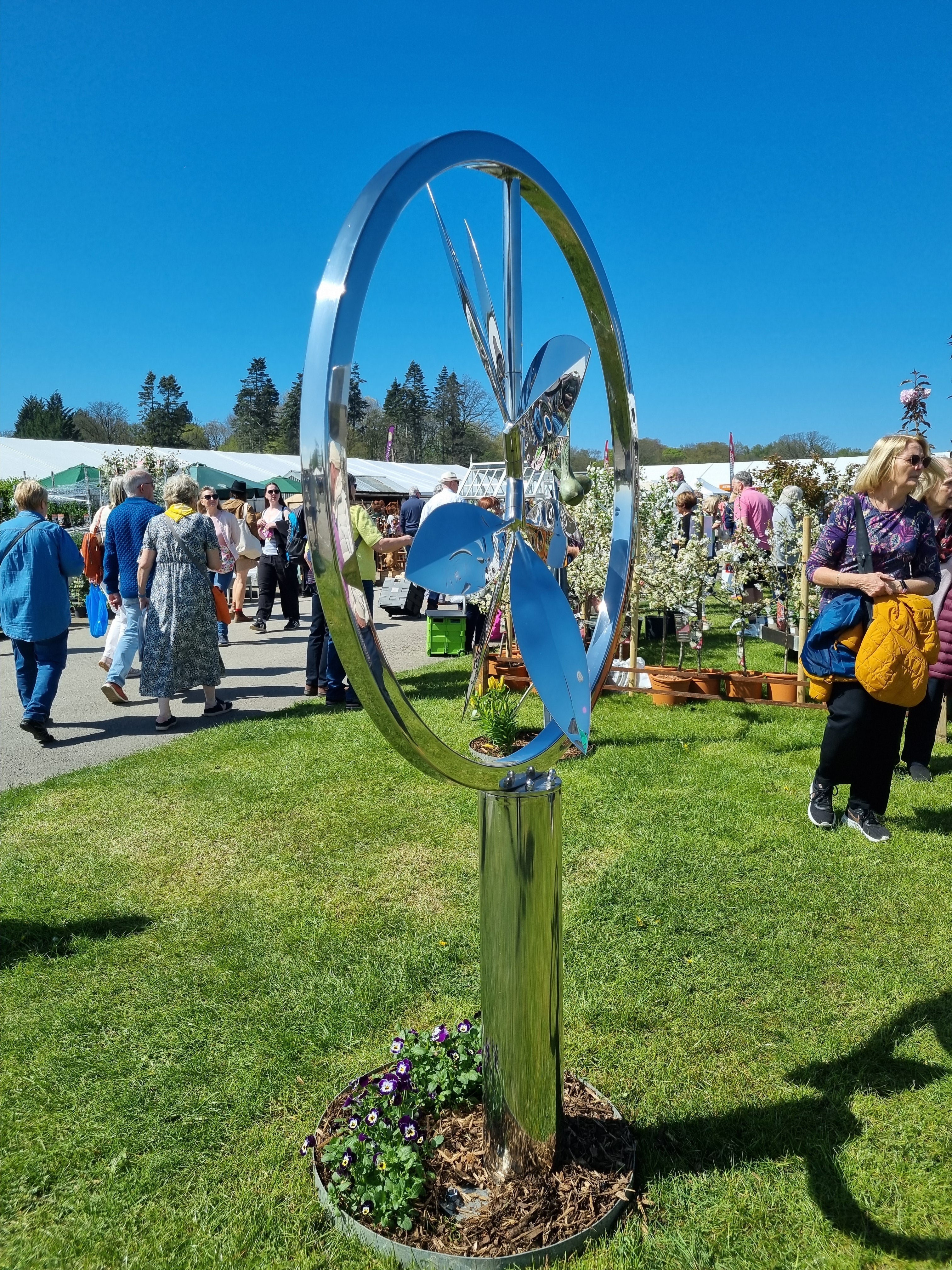 A reflective wind sculpture displayed at a garden show with visitors and gazebos visible in the background.