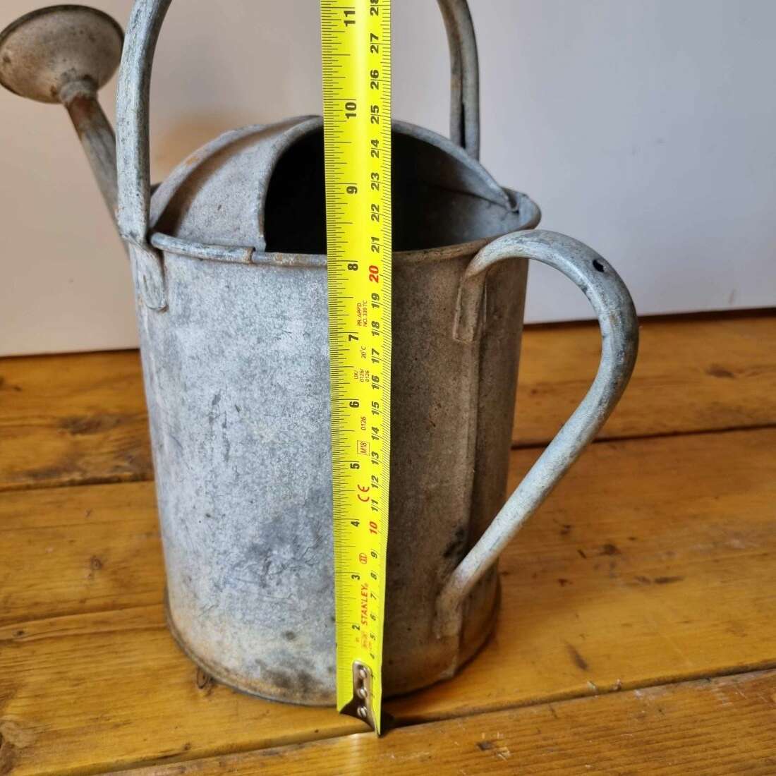 Vintage metal watering can with a measuring tape for scale on a wooden surface.