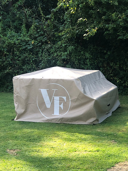 Beige outdoor furniture cover with 'VF' logo on a grassy area with greenery in the background