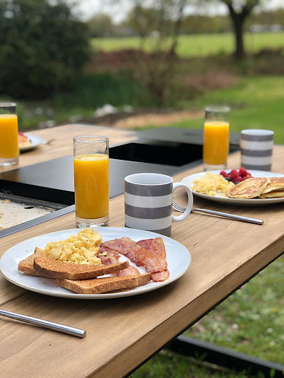 Breakfast scene with plates of food, glasses of orange juice, and a cup on a wooden table outdoors.
